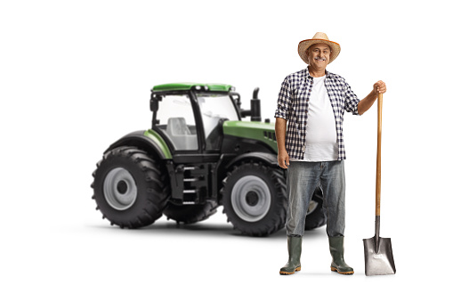 Mature farmer with a shovel standing next to a green tractor and smiling isolated on white background