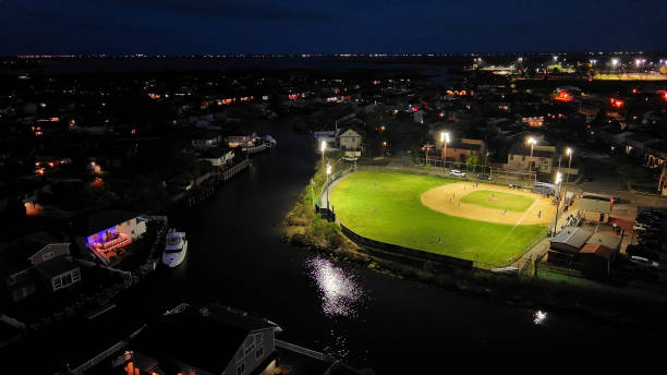Playing baseball at night. Amateur team playing baseball on illuminated public community stadium by sea channel at night. Oceanside, New York. Aerial view