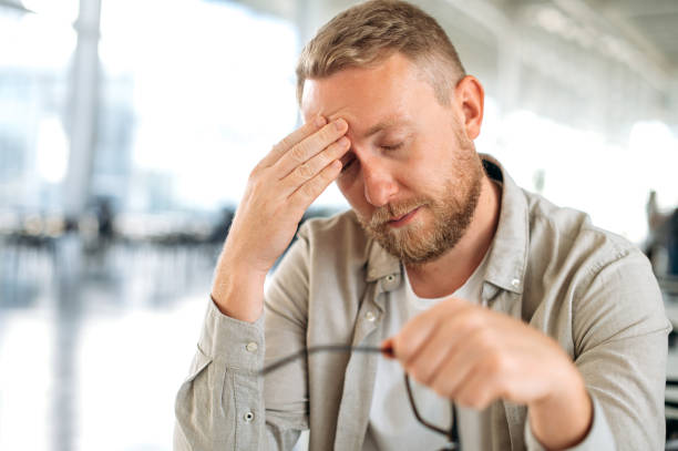 Exhausted caucasian man, company employee, IT specialist, sits at the workplace, took off his glasses, closed his eyes, tired from work, feels overtired, has a headache, need a rest or coffee brake stock photo