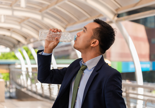 Side view of handsome Asian businessman in suit standing holding mineral water bottle and drinking for health and refresh in heat city. Portrait of happy young business drink pure water outside office.