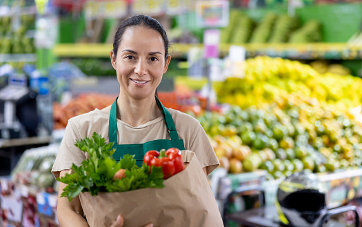Latin American saleswoman holding a bag of groceries while working at the supermarket and looking at the camera smiling