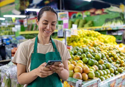 Happy Latin American retail clerk using her phone on her break while working at the supermarket