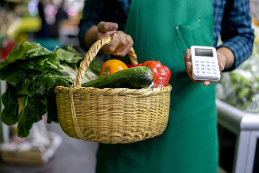 Close-up on a retail clerck holding a basket of food and a credit card reader while working at a greengrocer's shop