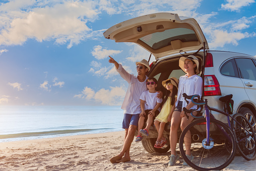 Happy Asian family with trip car travel having fun and enjoying on beach in summer vacation holiday. Portrait of father, mother, daughter, and son smiling cheerful in hatchback with bicycle on coast.