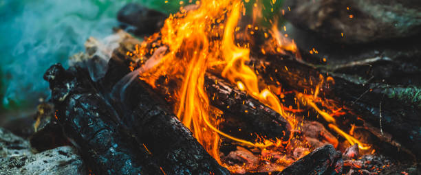 istock Vivid smoldered firewoods burned in fire close-up. Atmospheric background with orange flame of campfire and blue smoke. Warm full frame image of bonfire with glowing embers in air. Bright sparks bokeh 1487640956