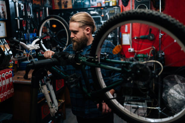 réparateur barbu réparant un vélo de montagne sur un support à vélo travaillant dans un atelier de réparation de vélos avec un intérieur sombre. mécanicien masculin effectuant des diagnostics - cycling bicycle occupation men photos et images de collection