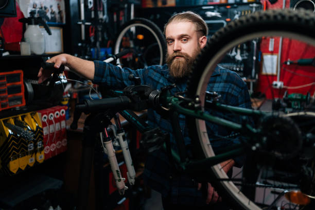 portrait d’un réparateur de vélos barbu debout derrière un vélo dans un atelier de réparation avec un intérieur sombre, regardant sérieusement la caméra. - cycling bicycle occupation men photos et images de collection