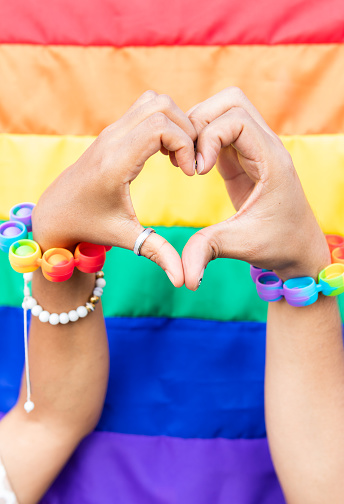 Two people's hands wearing pride bracelets, forming a heart shape against a rainbow flag background