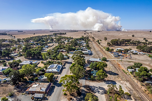 Aerial view of billowing smoke plume from multiple controlled crop stubble burnoffs near small rural farming town of Farrell Flat in mid-north of South Australia. In the foreground are houses and tree-lined streets plus an abandoned railway line heading north out of town. Burning crop stubble is a long-established farming practice that continues in Australia, despite moves to reduce the practice due to environmental and other concerns.