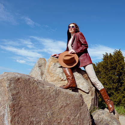 A lonely adult woman stands on the top of a cliff, looking into the vast expanses of the azure sky. A woman with long hair in a leather jacket stands on the rocks near the forest. Woman with sunglasses