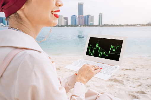 Young woman working on laptop with stock market exchange and cryptocurrency online graph and chart, against skyline of Abu Dhabi downtown city. Success and passive income concept