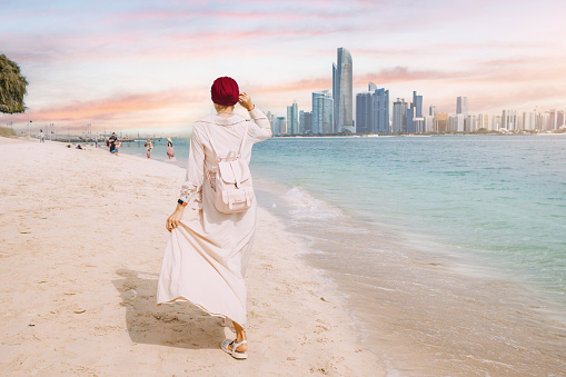 Despite the size and grandeur of the skyscrapers, the woman seems undaunted as she walks confidently along the shoreline, perhaps lost in thought or simply enjoying the peaceful moment.