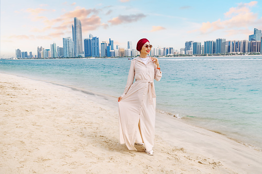 a young indian woman walks along the beach, taking in the breathtaking sea view of Abu Dhabi's towering skyscrapers.