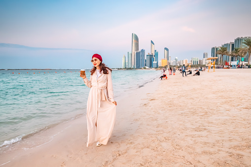 Happy girl walking on sandy beach with takeaway coffee with Abu Dhabi downtown skyscrapers in the background