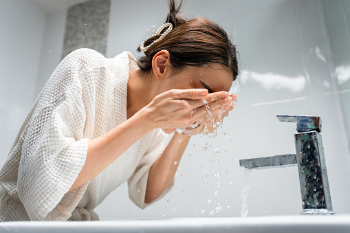 Asian beautiful woman washing her clean face with facial foam and water. Attractive female in bathrobe washing face for healthy beauty treatments and skin care then looking at the mirror in bathroom.