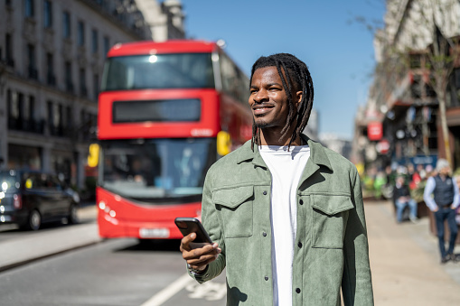 Waist up candid portrait of 31 year old black male walking on Regent Street holding smartphone, looking away,  red London double decker bus in background.