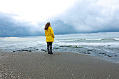 Nervous young woman in yellow raincoat standing alone by the sea on a rainy day