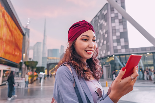 Happy asian girl using smartphone wireless network to connect to internet while walking in Dubai downtown with Burj Khalifa tower and other skyscrapers in background