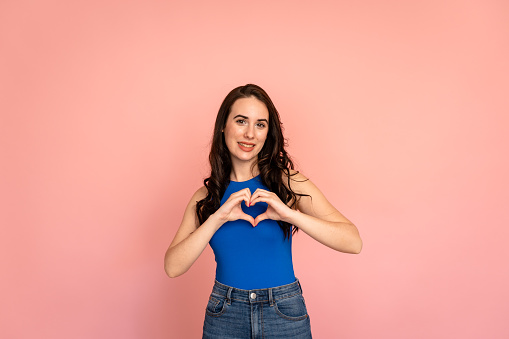 Woman making heart with her hands, showing positive feelings and love against pink background.