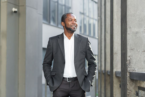 Portrait of a successful senior African-American businessman, lawyer, banker standing near a business center in a suit, holding his hands in his pockets, looking to the side with a smile.