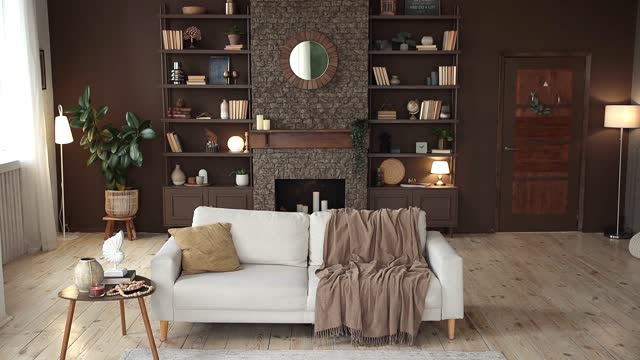 Modern stylish living room with large windows and beige sofa on the background of brown wall with fireplace, shelving with books and decor, and potted plants. Cozy chalet interior. Empty space