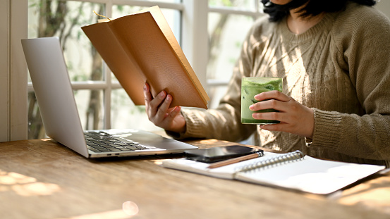 Cropped image of an Asian woman reading a book while enjoying with her iced matcha green tea at a beautiful minimal coffee shop.