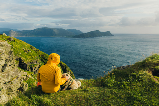 Happy male in yellow jacket sitting with a dog at the edge of the island looking at the islands and the ocean during summer sunset in Scandinavia