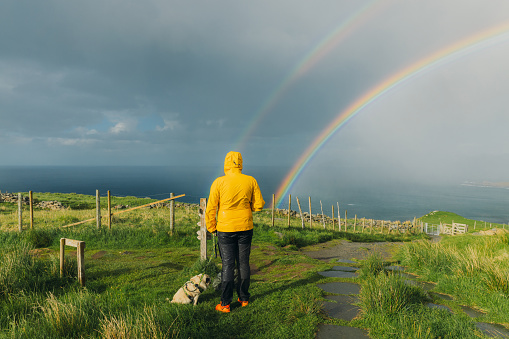 A male hiker in yellow jacket and his small pug admiring the hiking trip on green meadow with view of the ocean in Scandinavia