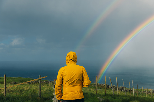 A male hiker in yellow jacket and his small pug admiring the hiking trip on green meadow with view of the ocean in Scandinavia