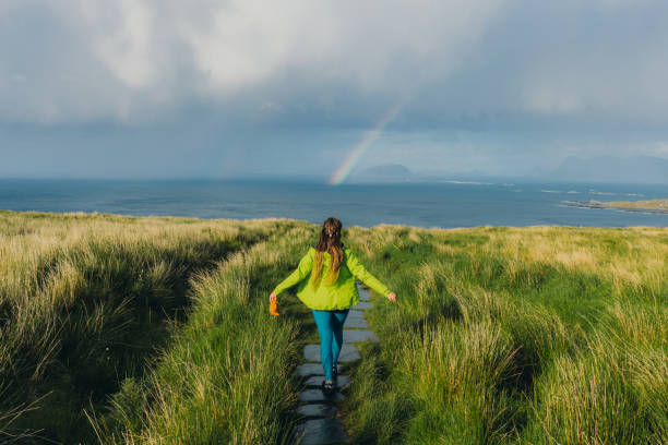 Happy woman hiking on island Runde under scenic rainbow from sea  in Norway stock photo