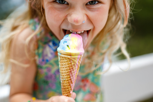 Happy preschool girl eating colorful ice cream in waffle cone on sunny summer day. Little toddler child eat icecream dessert. Sweet food on hot warm summertime days. Bright light, colorful ice-cream.