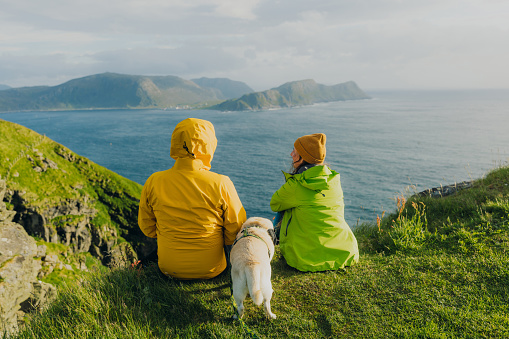 Heterosexual couple in yellow green jackets sitting with a dog at the edge of the island looking at the islands and the ocean during scenic summer sunset in Scandinavia