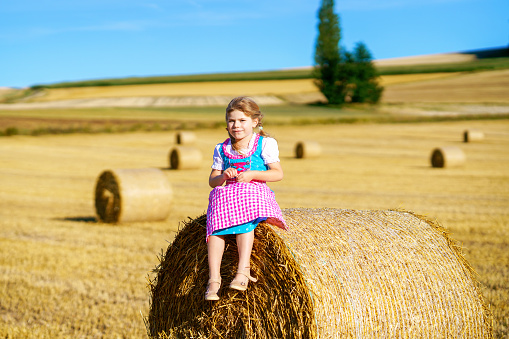 Cute little kid girl in traditional Bavarian costume in wheat field. Happy child with hay bale during Beer Fest in Munich. Preschool girl play at hay bales during summer harvest time in Germany