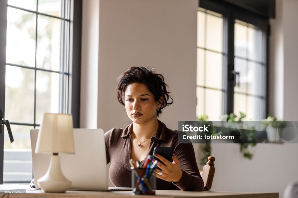 Young woman working at her desk at the office, using laptop Portrait of diligent young woman sitting at her office desk, using laptop and reaching for her smart phone, checking notifications and text messages. Text Messaging Stock Photo