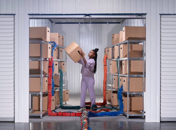 Cloud computing or storage is often depicted with ethereal images of stylised clouds. Yet, these technologies are not immaterial nor endless. This photo is a new take on the physical reality of these technologies and their environmental impact. mujer de pie en un garaje de paredes blancas con estanterías y cajas de almacenamiento de cartón, sosteniendo una caja. los paquetes de cables ethernet corren hacia el garaje entre las estanterías. una representación simbólica de 'cloud computing' o '