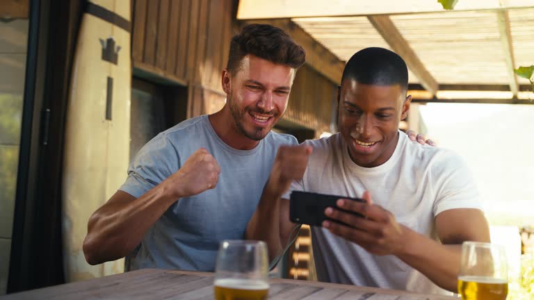 Two Male Friends Outdoors At Home Watching Sports On Mobile Phone Celebrating Team Win