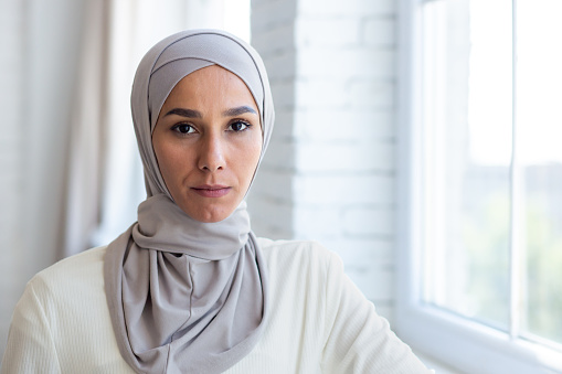 Close-up photo. Portrait of a young Muslim woman in a hijab standing by the window at home, confidently and seriously looking at the camera.