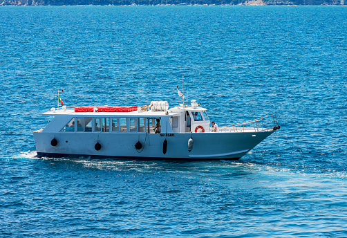 Tellaro, Italy - July 9th, 2022: White ferry boat with a small group of tourists, in motion in the blue Mediterranean sea (Ligurian sea) in front of the ancient and famous village of Tellaro on a sunny summer day, tourist resort on the coast of the Gulf of La Spezia or Gulf of Poets, Lerici municipality, Liguria, Italy, southern Europe.