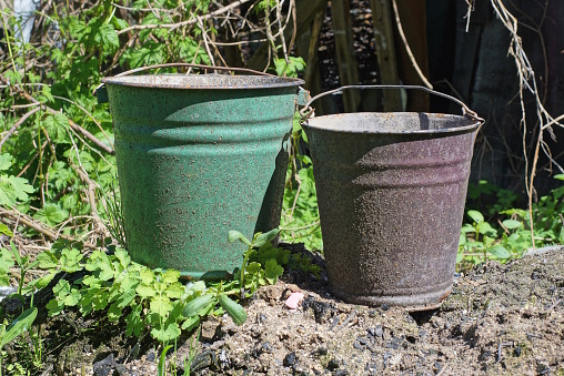 two old dirty metal enameled buckets stand on the gray earth among the green vegetation on the street