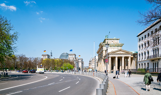 The photo captures the beauty of Berlin in a sunny spring day, with people strolling near the iconic Brandenburger Tor in the Mitte district. The Deutscher Bundestag can be seen in the background, adding to the grandeur of the image. The side view of the Brandenburger Tor and Deutscher Bundestag offers a unique and beautiful perspective of these iconic landmarks.
