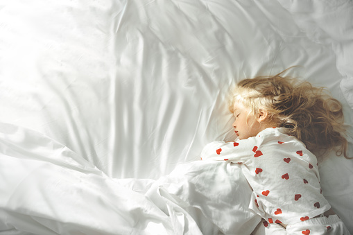 Cute little blonde girl in white pajamas with red hearts sleeping deeply on white bed. copy space, Top view