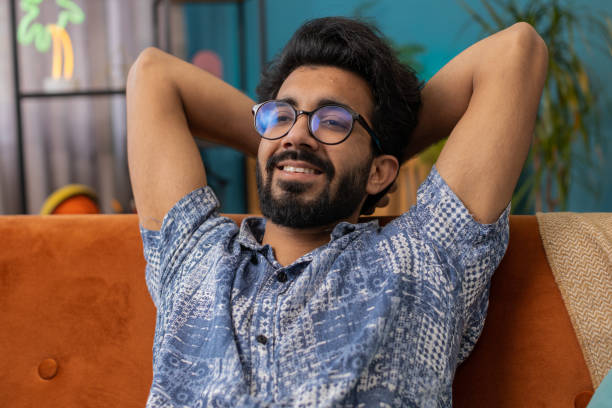 Tired indian man lying down on comfortable couch, taking a rest, daytime nap. Portrait of carefree hindu guy holding hands behind head relaxing, falling asleep at home apartment living room on sofa