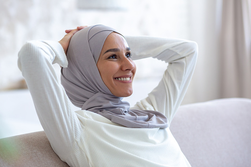 Close-up photo. Smiling arab young business woman in hijab sitting on sofa at home with hands behind head and resting, smiling and satisfied with successful work done and finished.