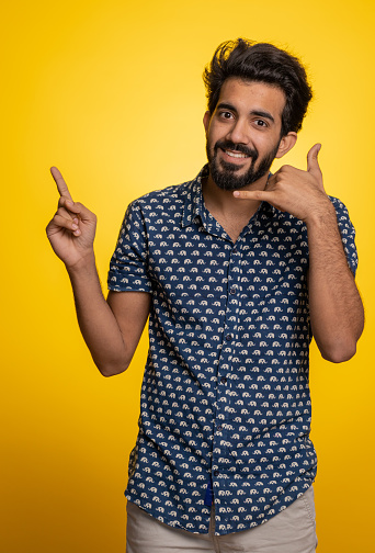 Call us, here is contact number. Handsome bearded indian man in shirt looking at camera doing phone gesture like says hey you call me back conversation. Hindu guy isolated on yellow background, indoor