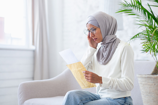 Muslim woman at home in hijab upset, arab woman received mail message notification with bad news, woman reading pensive and worried sitting on sofa in living room.