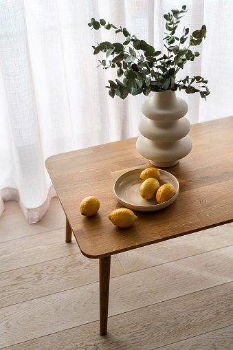 Still life composition with lemons in bowl close to white vase with eucalyptus plants. Floral decor on wooden table in kitchen or living room, vertical shot