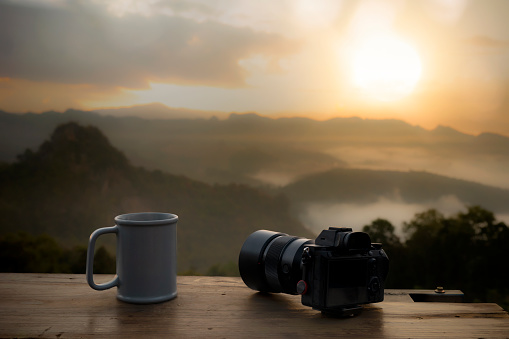 Close-up of camera and coffee cup on wooden table in the morning with mountain and nature background