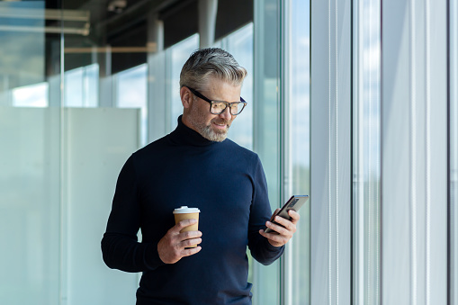 Senior male businessman stands in the office center by the window and uses the phone, writes messages, reads news, mail. He holds a cup of coffee in his hand. Coffee break, rest, waiting to meet.