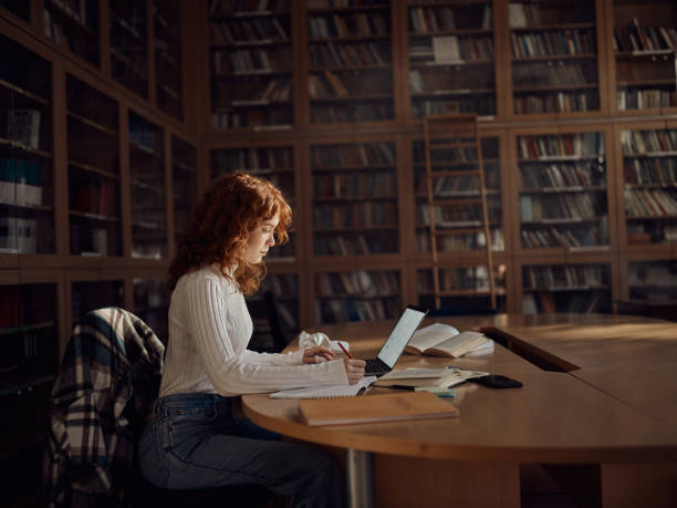 estudiante femenina tomando notas mientras e-learning en la biblioteca. - book young adult women student fotografías e imágenes de stock