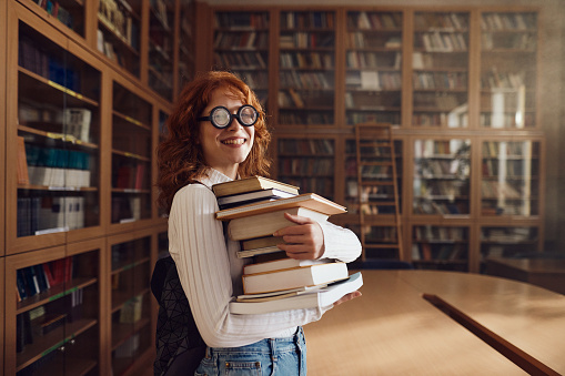 Happy female student holding stack of books in library and looking at camera. Copy space.